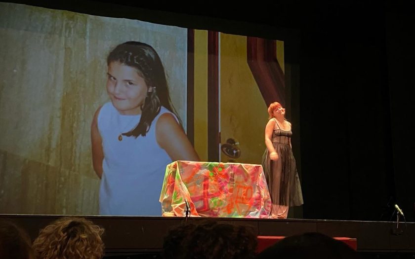 Encenação teatral no escuro com uma atriz no cenário e no fundo imagem projetada duma criança com a mesma atitude e posicionamento