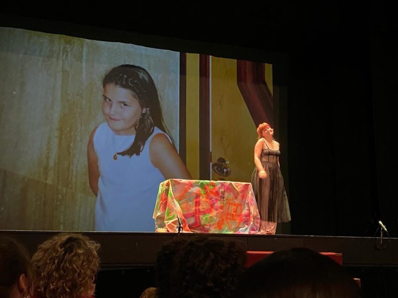 Encenação teatral no escuro com uma atriz no cenário e no fundo imagem projetada duma criança com a mesma atitude e posicionamento