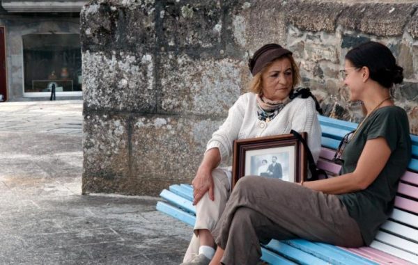 duas mulheres a falar num banco de cor azul ciano, à frente dum muro de pedra de granito. Entre elas uma grande fotografia num quadro
