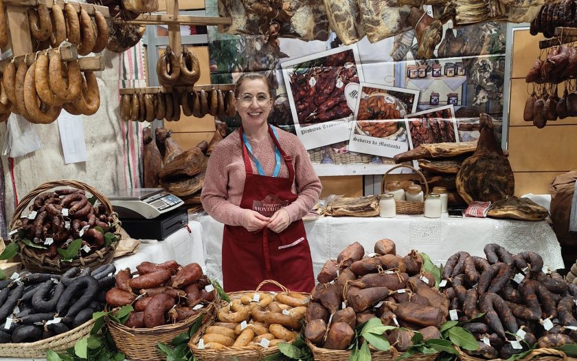 Posto de venda na feira carregado de produtos de fumeiro com vendedora a sorrir no centro