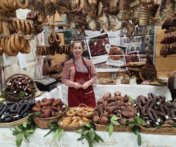 Posto de venda na feira carregado de produtos de fumeiro com vendedora a sorrir no centro