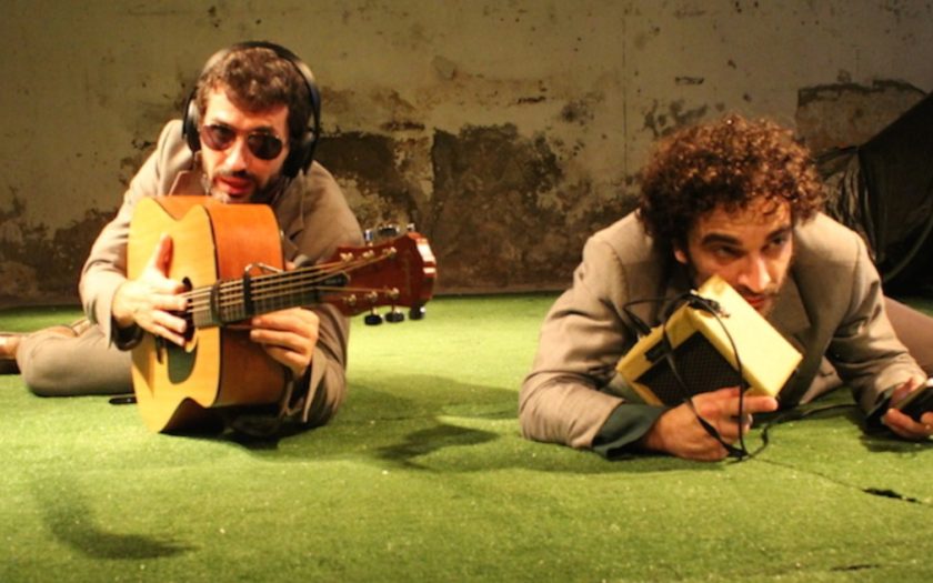 imagem de atuação com dois homens deitados num chão de cor verde com guitarras