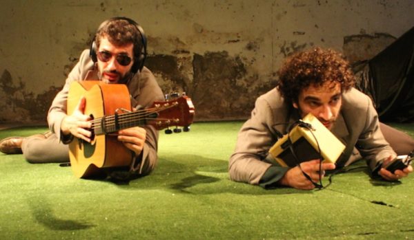 imagem de atuação com dois homens deitados num chão de cor verde com guitarras
