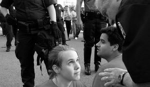 Rapaz jovem sentado na rua em protesto a olhar para um polícia que está a ralhar com ele